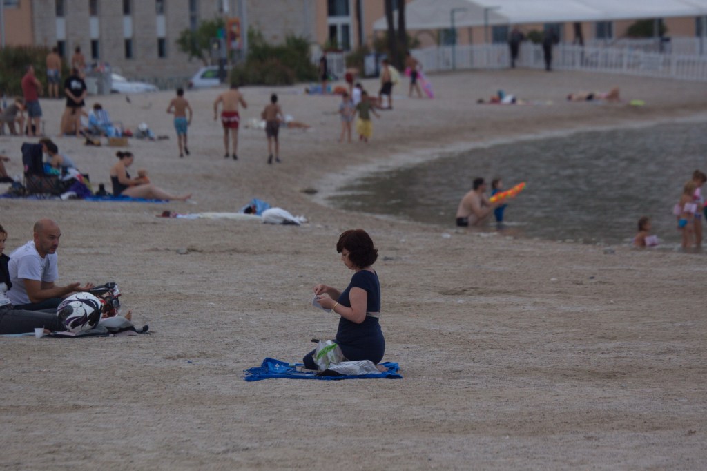 A woman sitting on a towel at the beach, enjoying a snack, while people are relaxing and playing in the background.