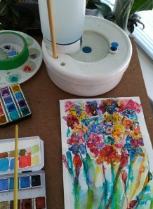 An artist's workspace featuring watercolor supplies and a vibrant floral painting in progress, showcasing a variety of colors and brushstrokes.