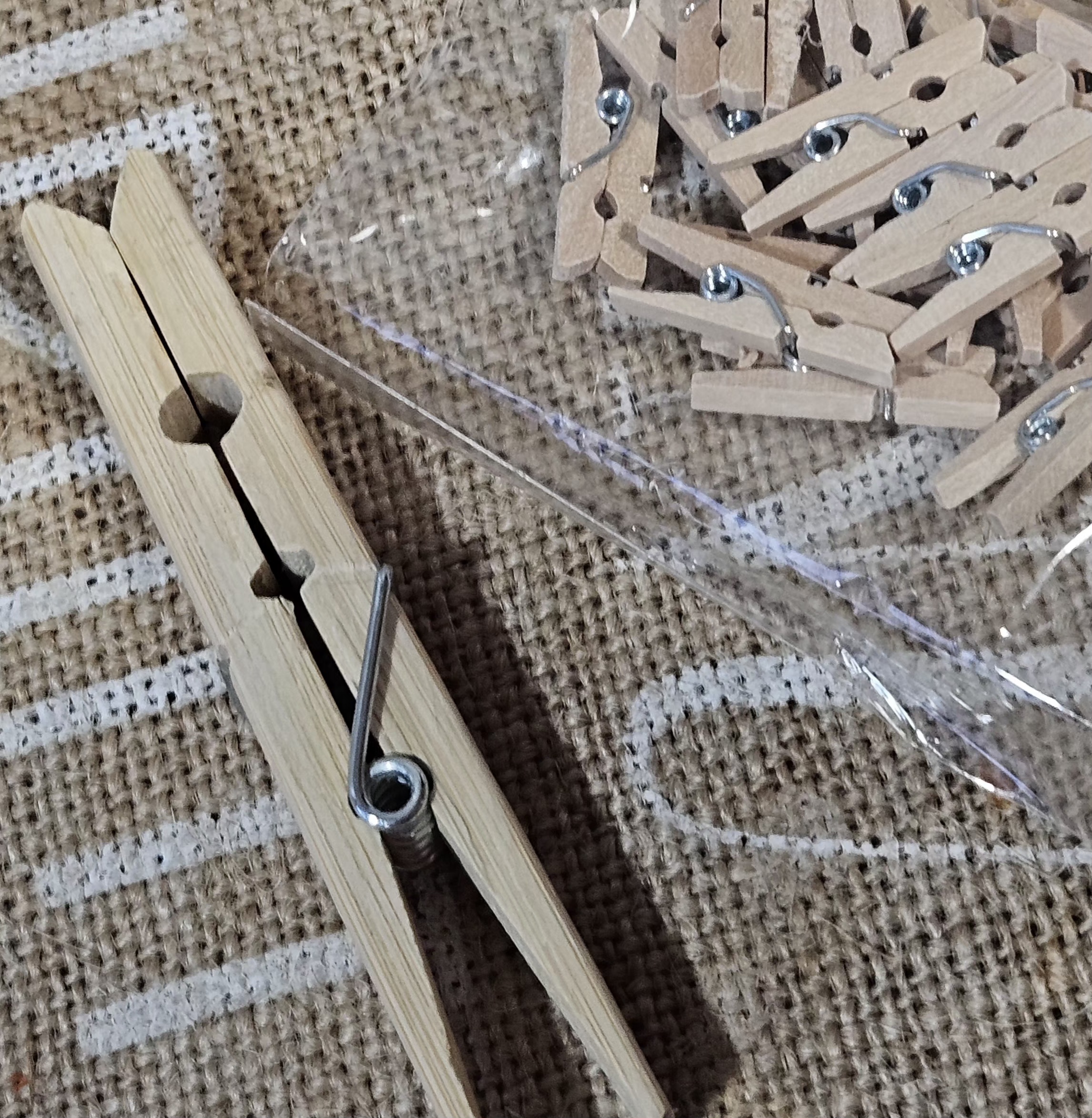 A clear container filled with small wooden clothespins on a textured fabric surface with two larger clothespins beside it